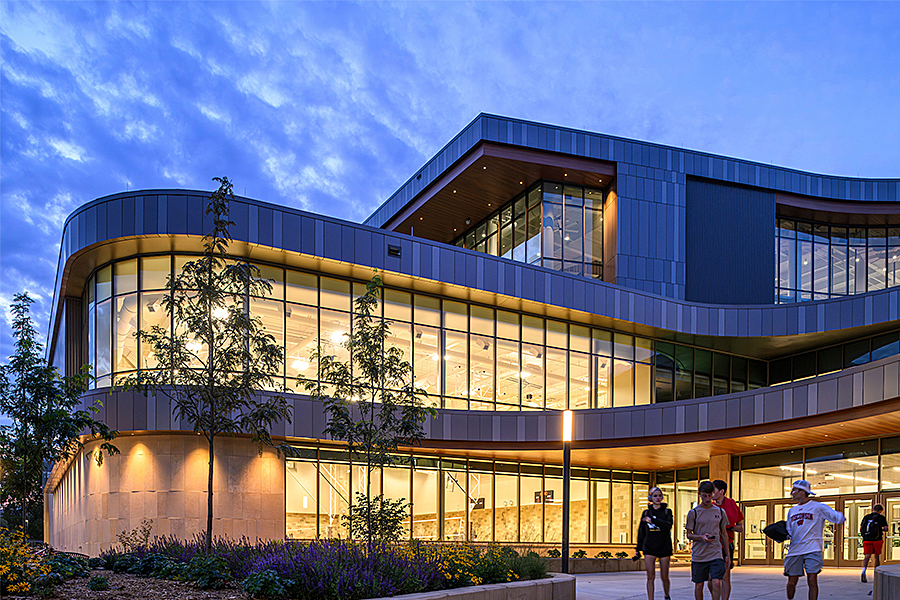 Glass exterior of a building glowing during evening with people walking in foreground. 