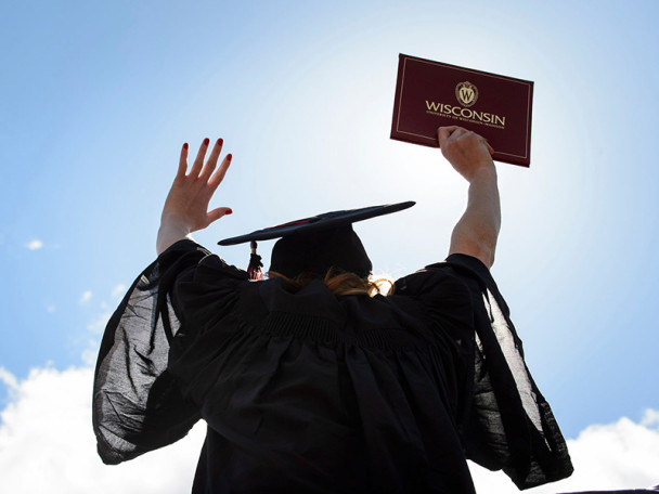 A student wearing a graduation robe and cap celebrates outdoors, lifting her diploma up in the air.